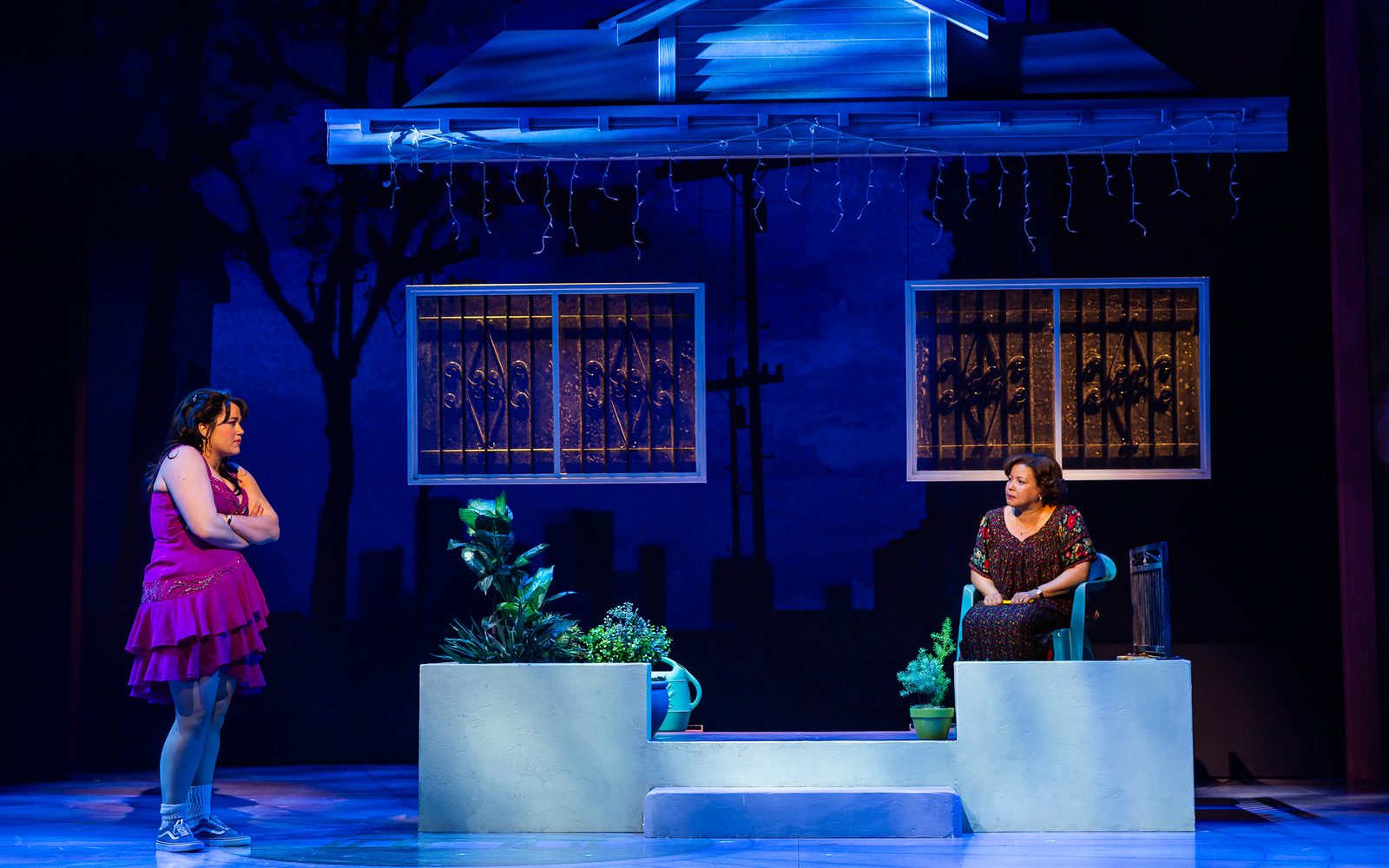 Lucy Godínez stands with her arms crossed in a pink dress, staring at Justina Machado sitting on the porch of their home in the dark.