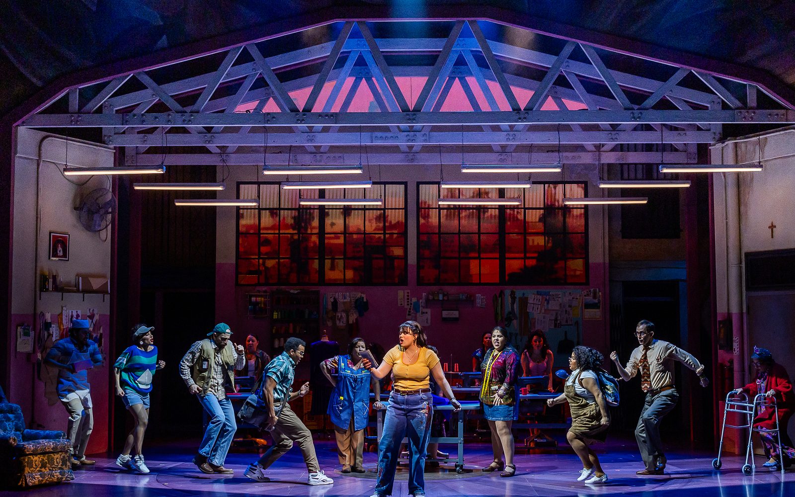 Lucy Godínez sings with a book in her hand while members of the cast gather onstage behind her in the garment factory.
