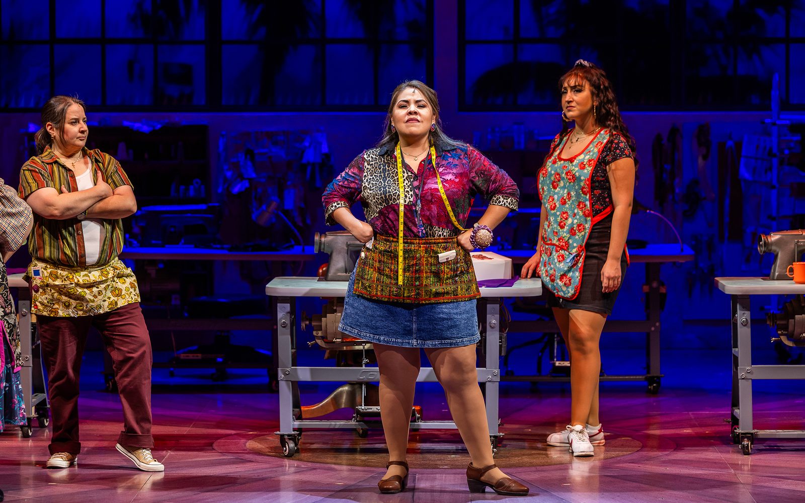 Florencia Cuenca in her sewers uniform stands proudly, with Sandra Valls and Shelby Acosta looking on from behind in the garment factor.