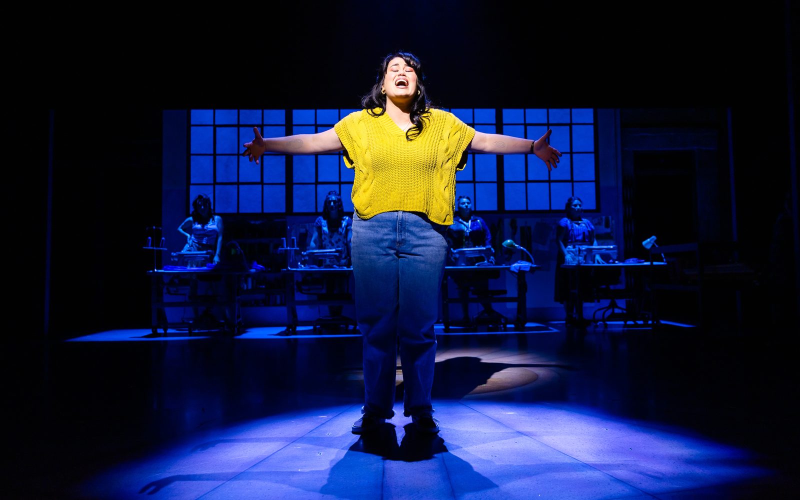 Lucy Godínez stands center with her arms flung open in a knit yellow top and lit brightly, while other cast members stand behind her at tables in dim blue lighting.