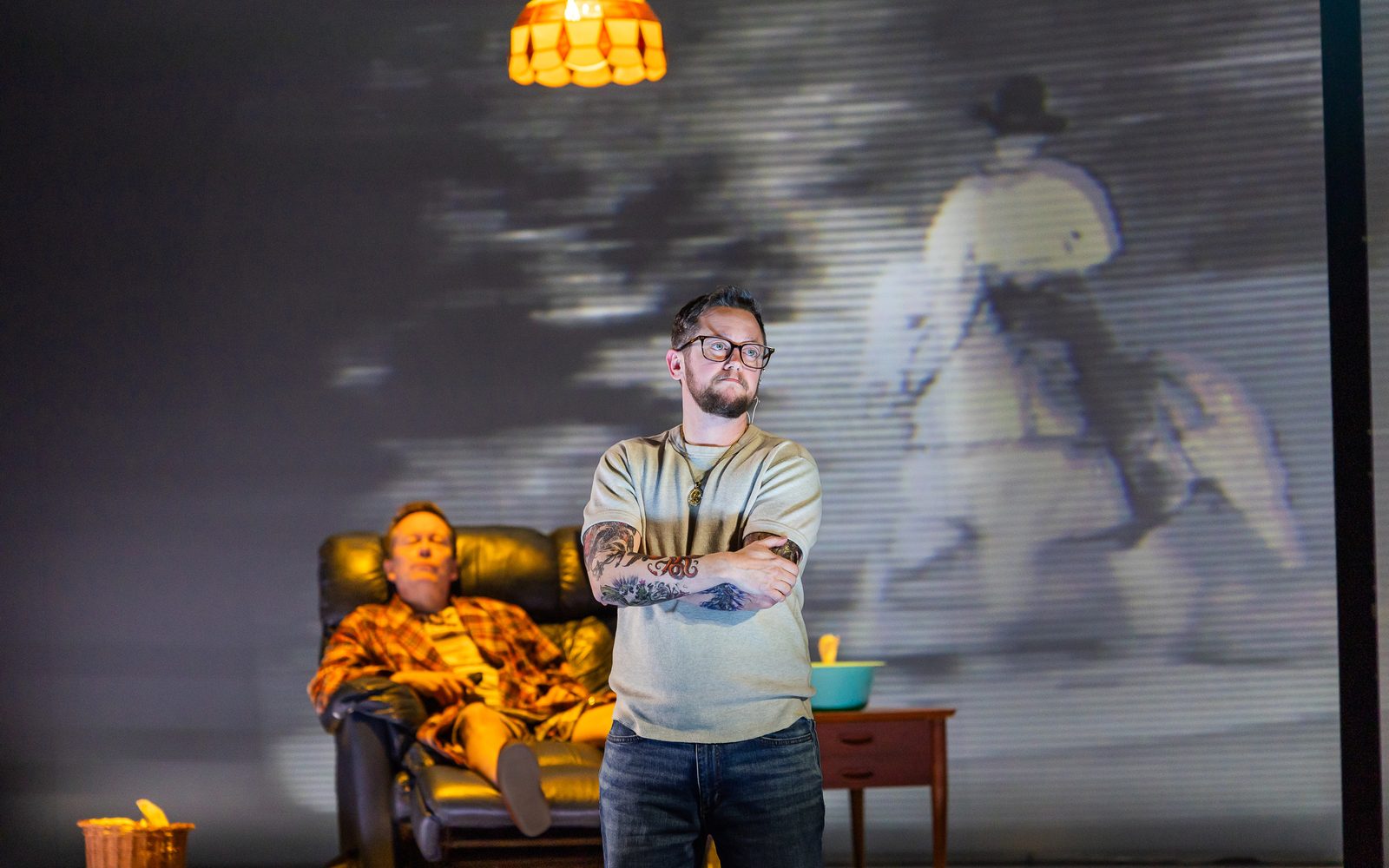 Christopher Liam Moore is bathed in yellow light seated in a recliner with Petey Gibson in front of him clutching his arms.