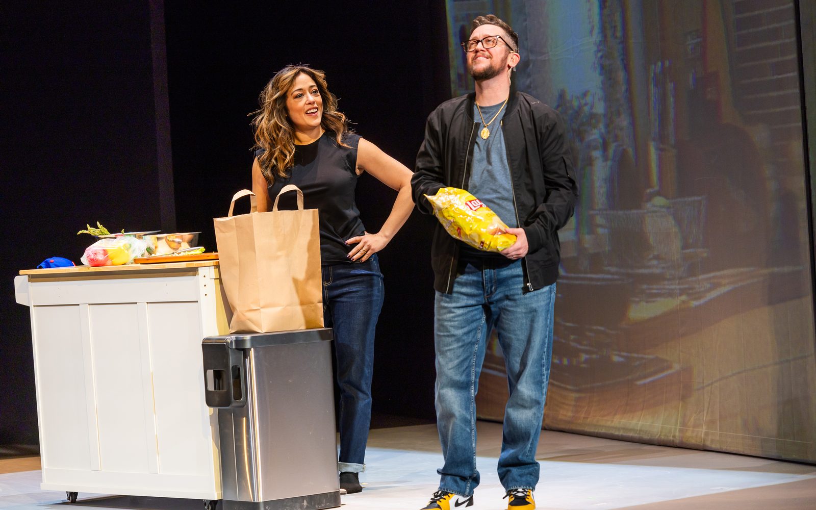 Elena Hurts leans on a kitchen island while Petey Gibson smiles while eating from a bag of potato chips.