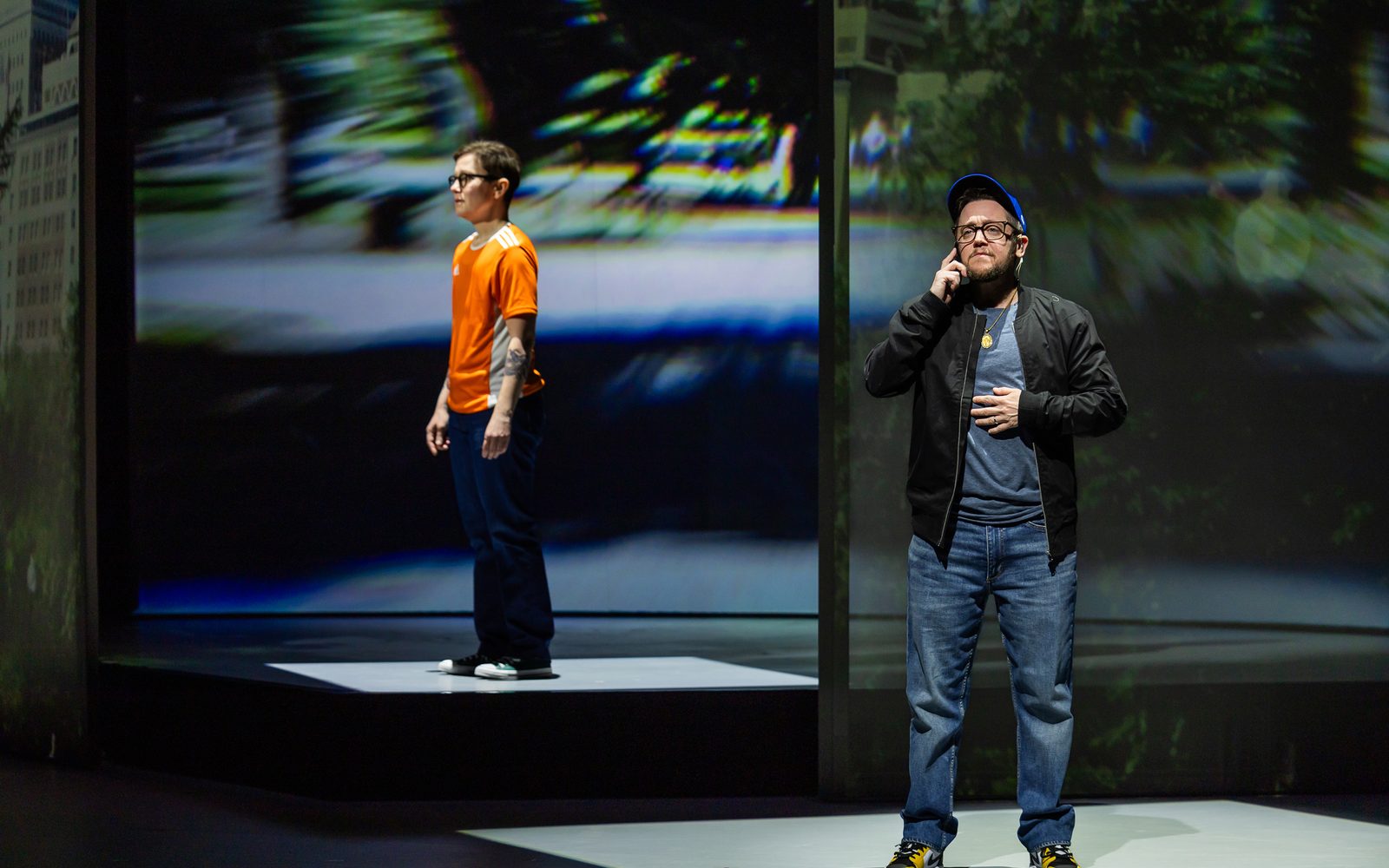 Stacey Raymond in an orange athletic shirt and Petey Gibson in a black jacket and blue cap stand apart, facing different directions. Petey has a phone up to his ear.