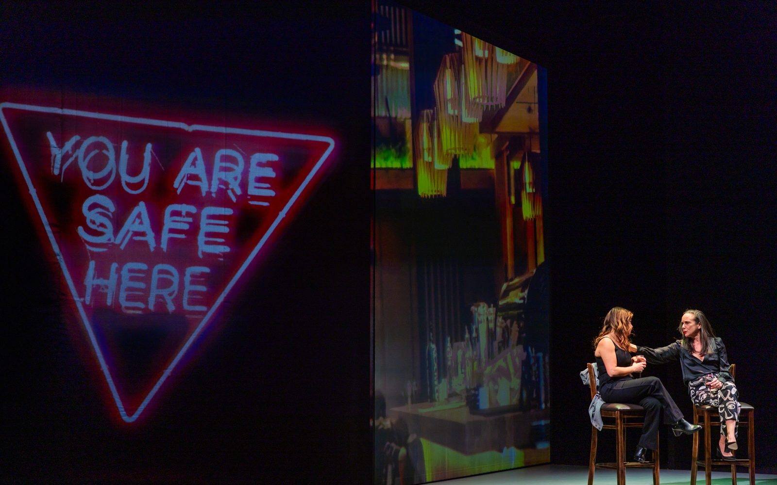Elena Hurst and Susan Rome sit on barstools. Susan reaches out to Elena's shoulder. A large sign says “You Are Safe Here” on the left.