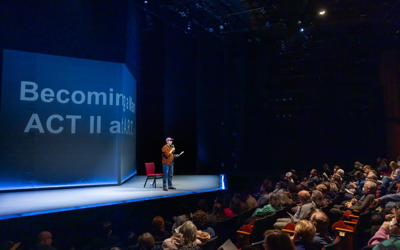 Lyam B. Gabel standing onstage speaks into a microphone as the audience listens.