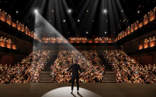 Looking out from the West Stage, with a performer spotlit onstage, people sitting in the orchestra level, boxes on either side, and a balcony.