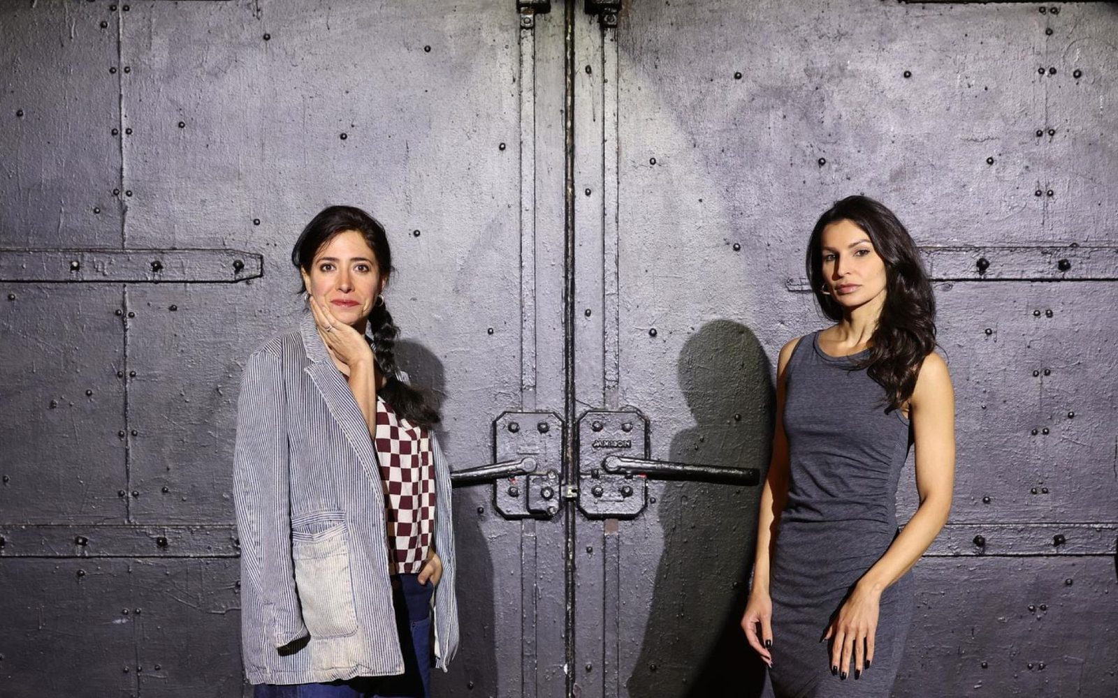 Rachel Chavkin with her hand on her chin and Martyna Majok with her arms at her side stand facing each other in front of large, black industrial doors.