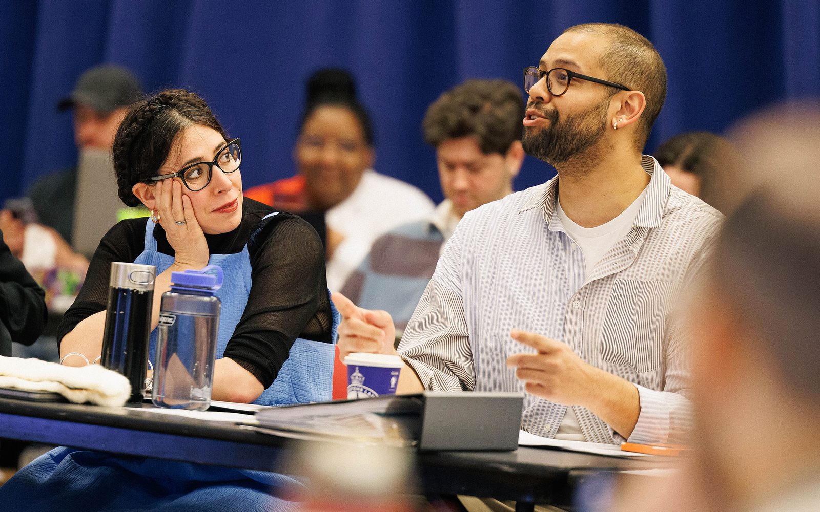 Seated at a table, Rachel Chavkin rests her head on her hand as she turns to look at Keenan Tyler Oliphant speaking next to her.