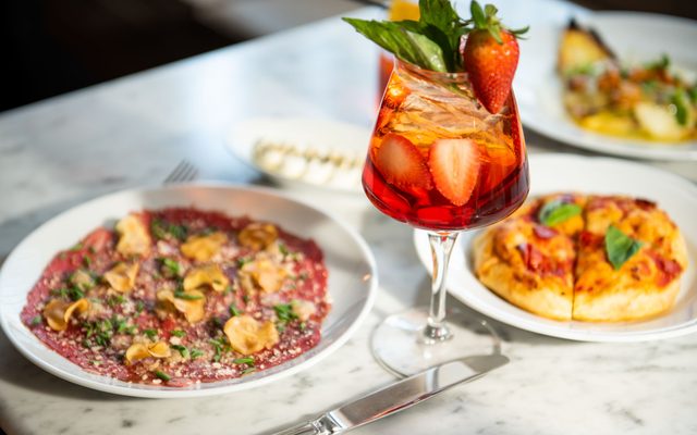 A glass of strawberry-filled sangria next to a plate of carpaccio and a miniature pizza.