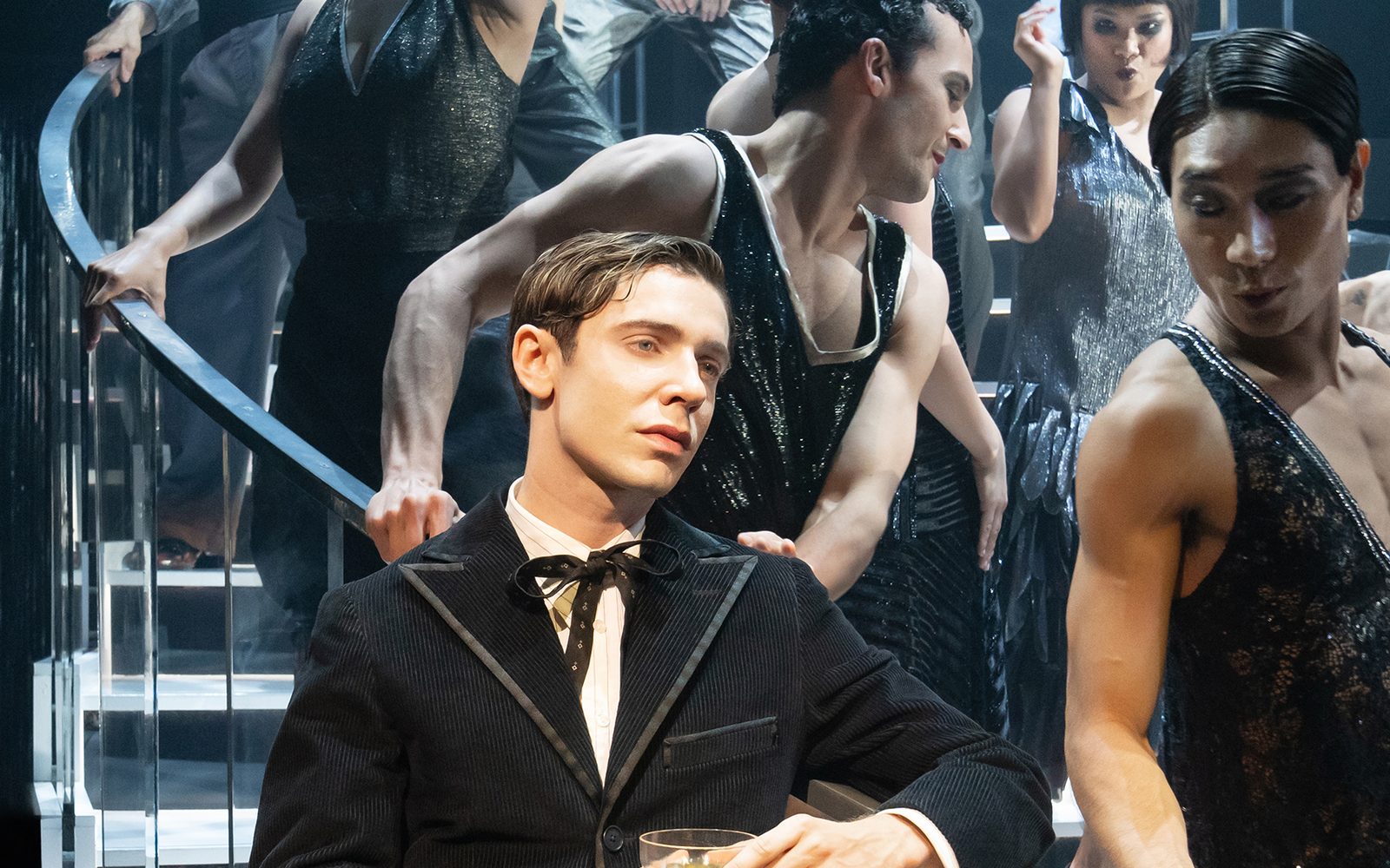 Ben Levi Ross in a black suit leans on a stair railing holding a coupe glass, while ensemble members dance down the stairs.