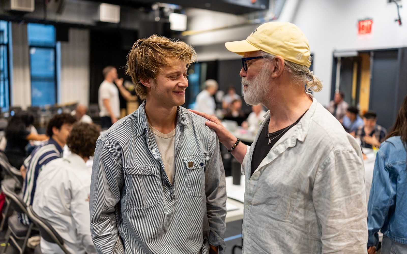 Rudy Pankow in a striped blue shirt smiles at Terrence Mann in a yellow baseball cap, who places his hand on Rudy's shoulder.
