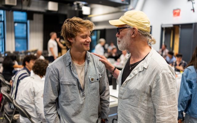 Rudy Pankow in a striped blue shirt smiles at Terrence Mann in a yellow baseball cap, who places his hand on Rudy's shoulder.
