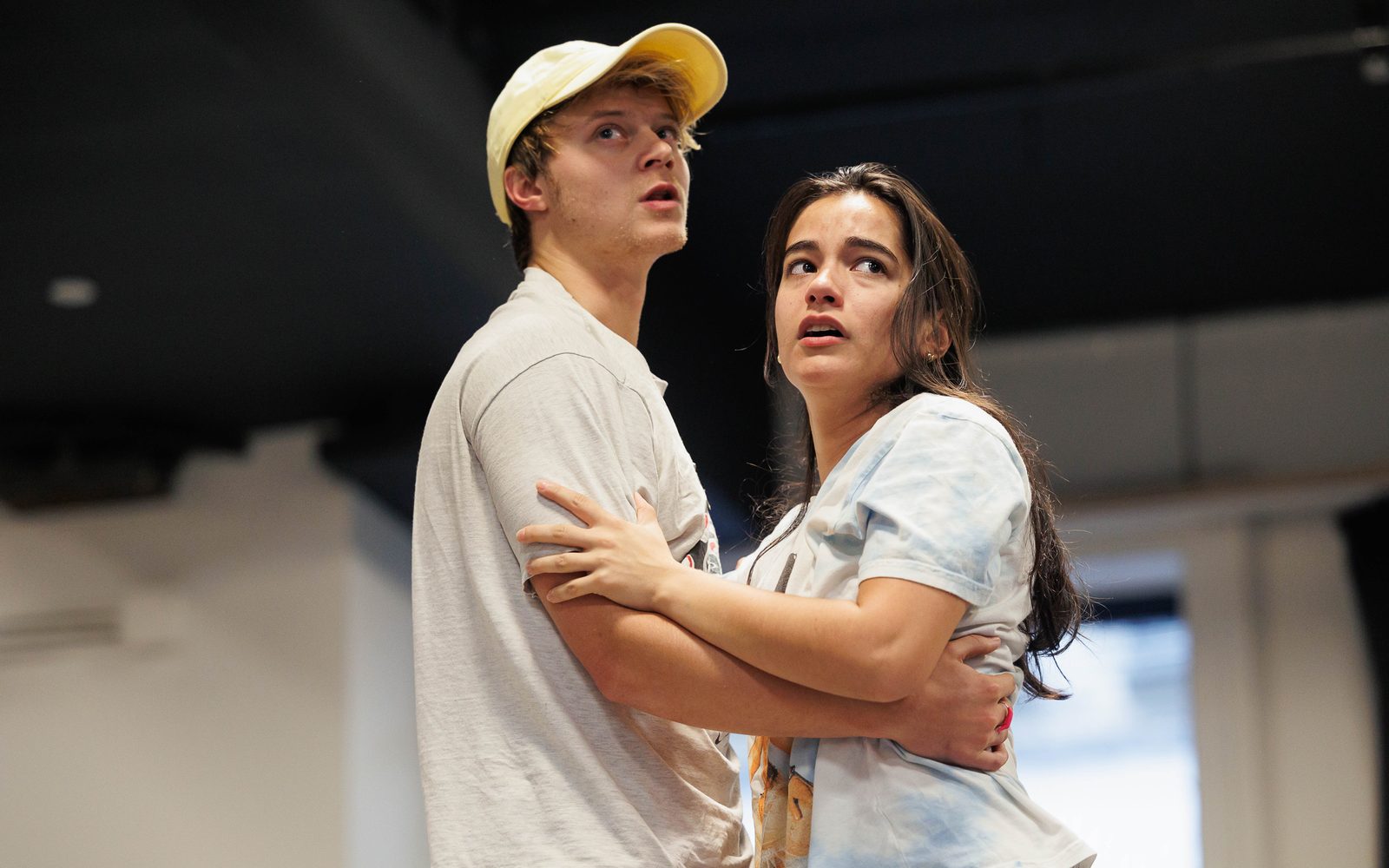 Rudy Pankow in a cream shirt and a yellow baseball cap and Emilia Suárez in a faded tie-dye shirt hold each other and look fearfully into the distance.