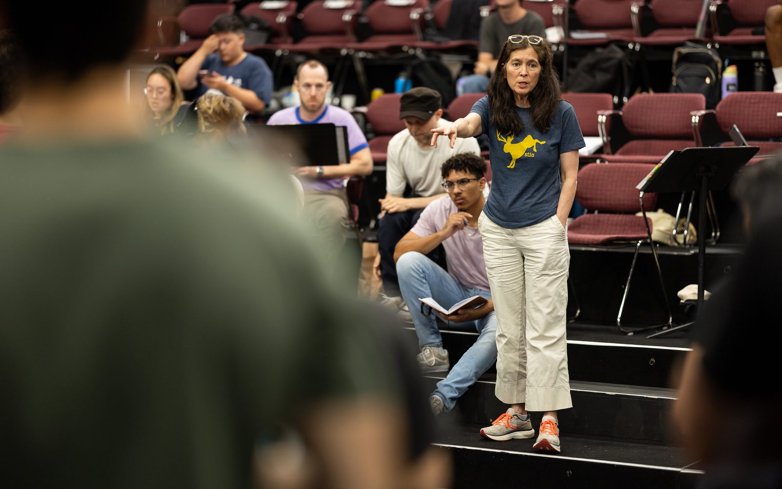 Diane Paulus, in a blue graphic T-shirt, stands on a raised platform gesturing to the stage, with other members of the creative team seated behind her.