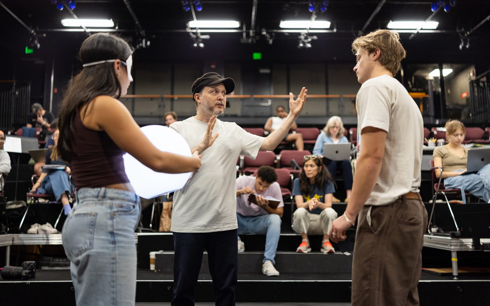 Sidi Larbi Cherkaoui in a black cap gestures to Rudy Pankow, on the right, and Emilia Suaréz, on the left holding a glowing orb.