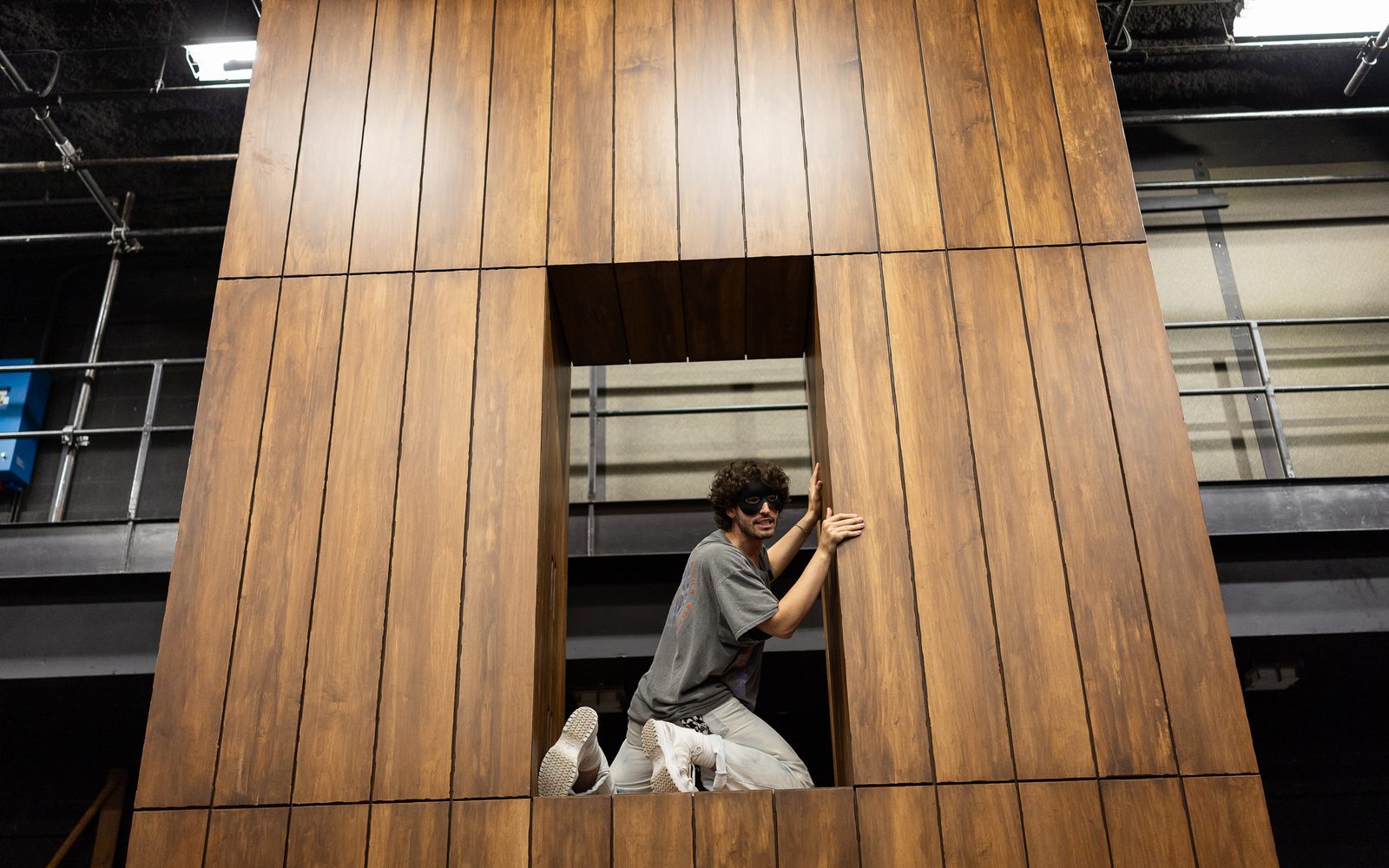 Clay Singer in a black half-face mask kneels in a portal in a wooden wall, turning to face out.