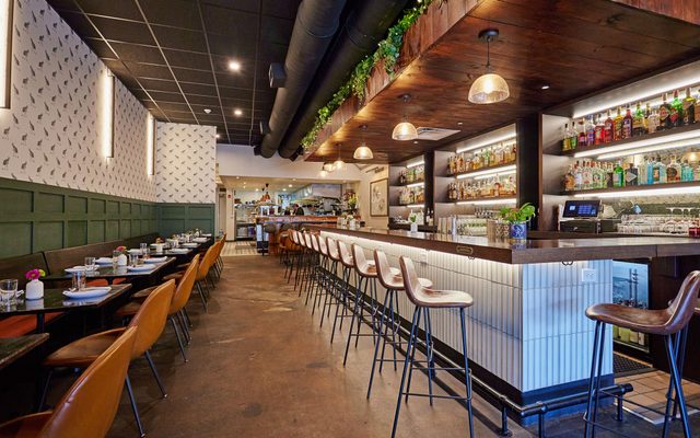 The interior of Season To Taste, with a wood-topped bar with high stools on the right, and tables with chairs on the left.