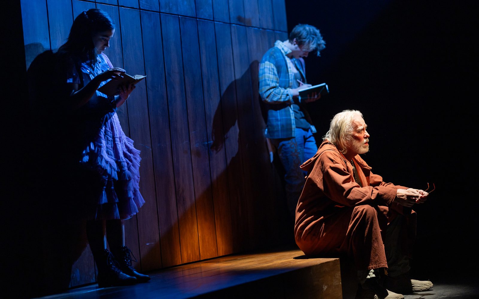 Terrence Mann in a maroon tunic is light brightly, while on either side of him lit in blue and leaning against a wall are Emilia Suárez reading a book and Rudy Pankow writing in a notebook.