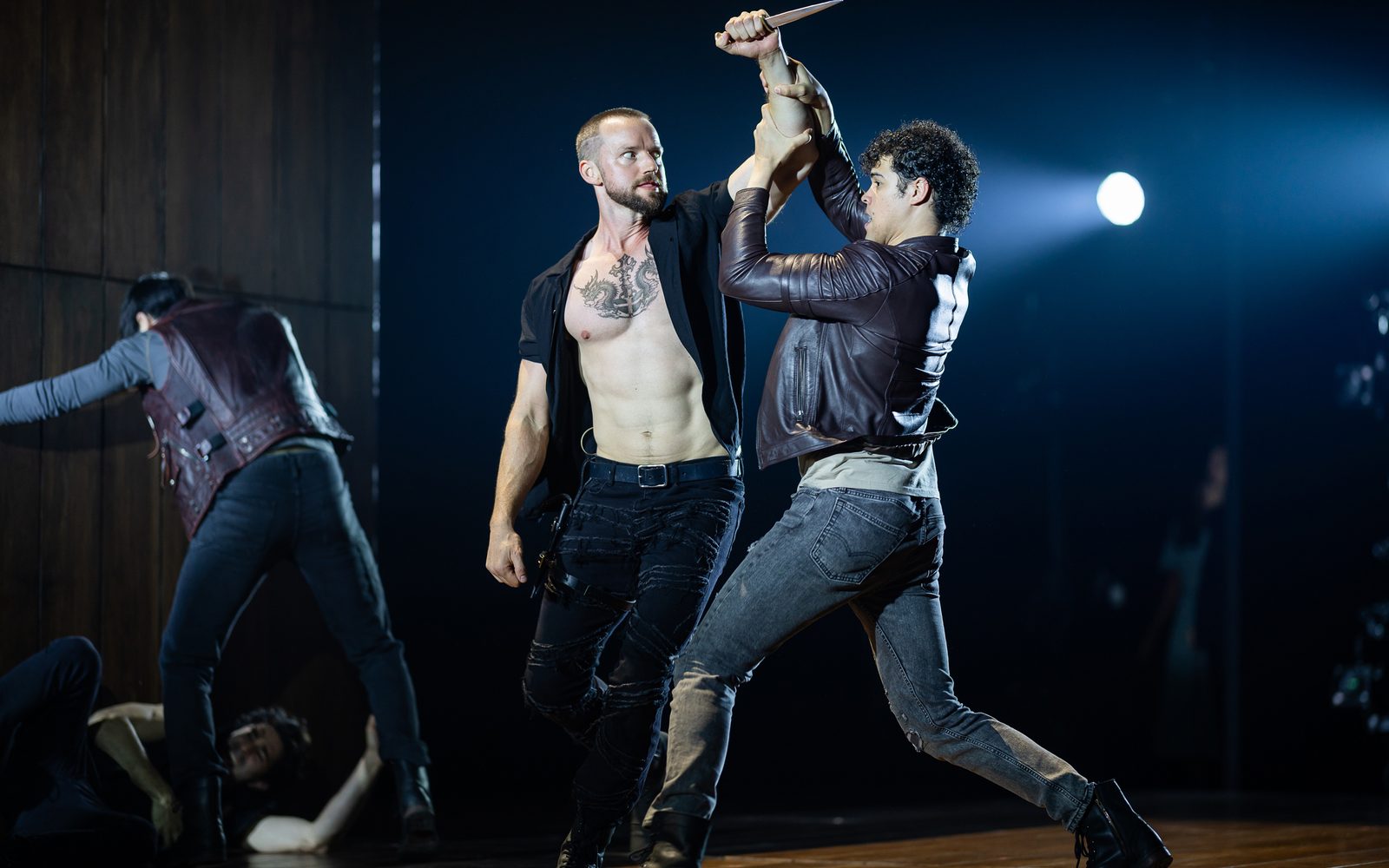 An angry-looking Alex Ross in an unbuttoned, short-sleeve black shirt raises a dagger above Brandon Dial’s head, who grabs his arm with both hands and a fearful look.