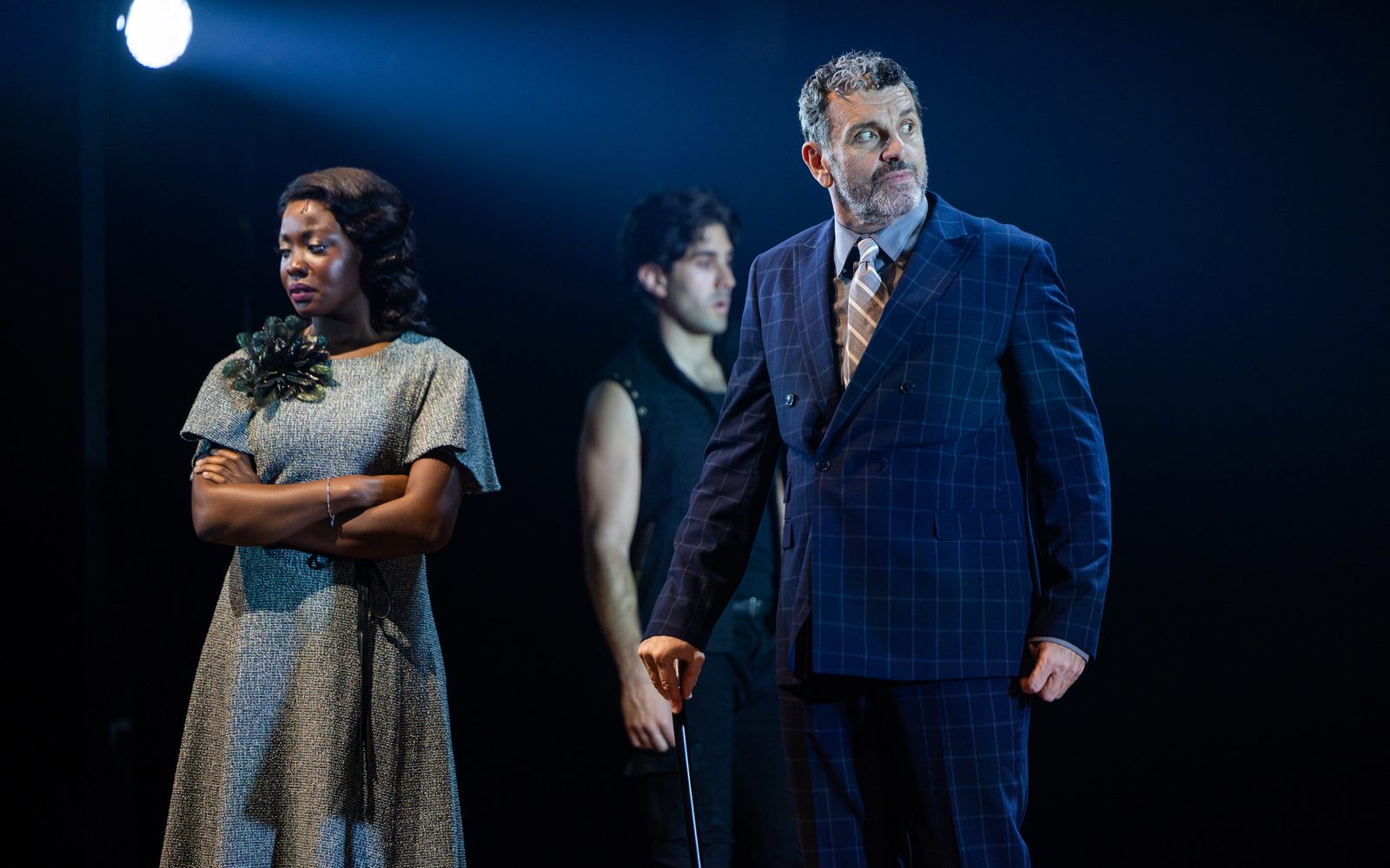 Abiola Obatolu in a grey dress folds her arms and looks downcast, while Bradley Dean, in a blue windowpane suit, looks warily to the side; Adam Shaukat walks behind them.