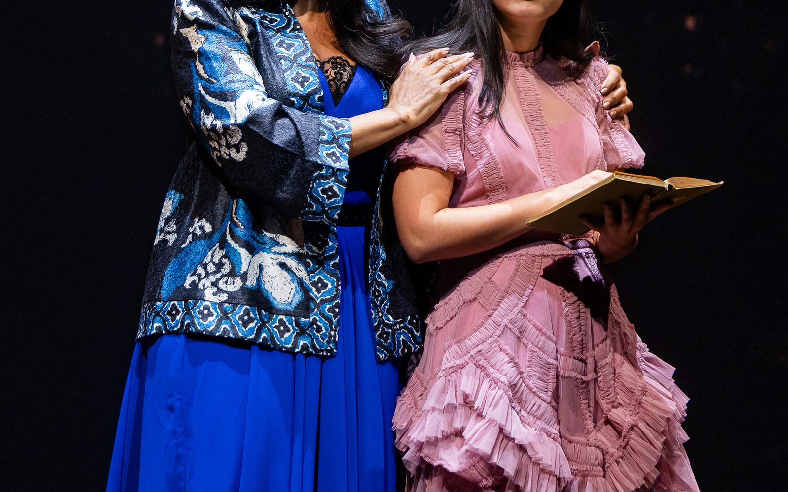 Sharon Catherine Brown in a blue dress and brightly patterned blue jacket places her hands on Emilia Suárez, in a pink dress with many frills and a book open in her hands.