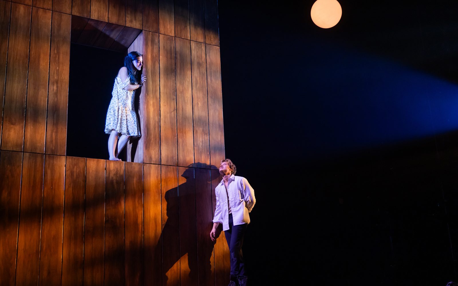 Emilia Suárez stands barefoot in a portal in a wooden wall, looking down at Rudy Pankow who looks up from below, while a yellow-orange moon hangs in the dark background.