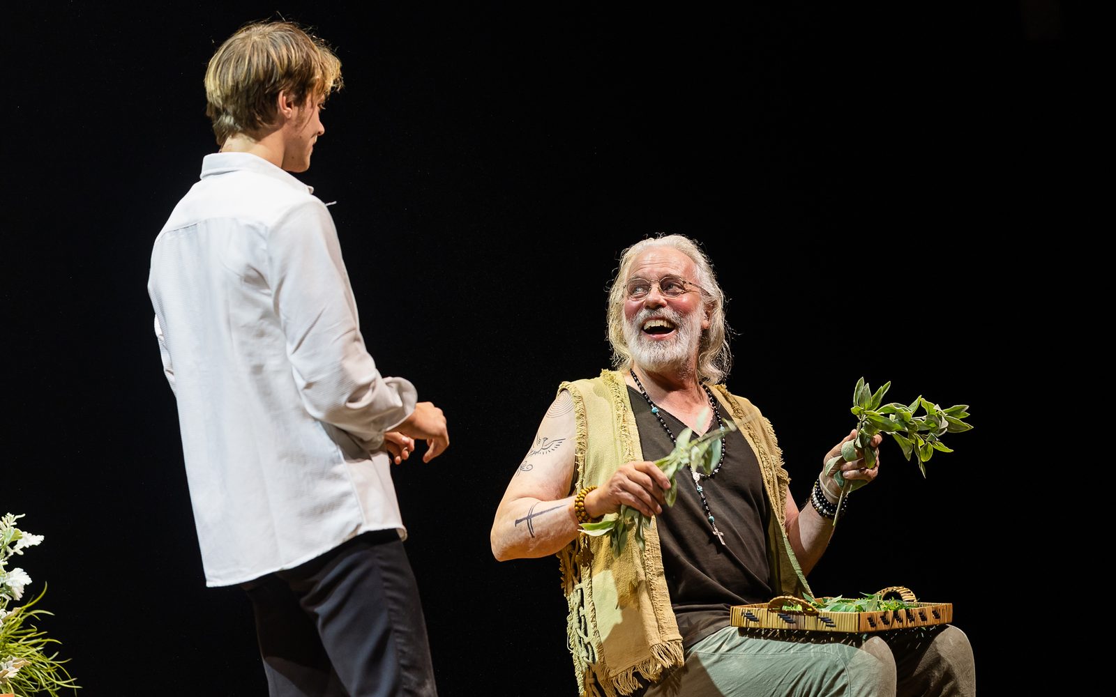 Rudy Pankow in a white dress shirt faces Terrence Mann, sitting and smiling broadly with herbs in his hand a small tray on his lap.