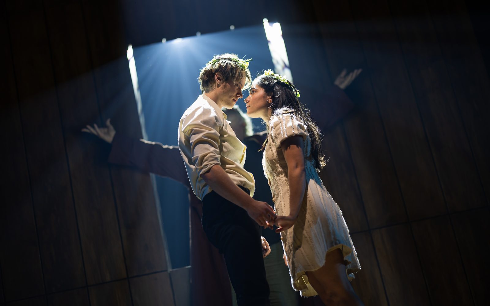 Light shines down on Rudy Pankow and Emilia Suárez, who stare at each other as if to kiss, each holding the other’s hand.