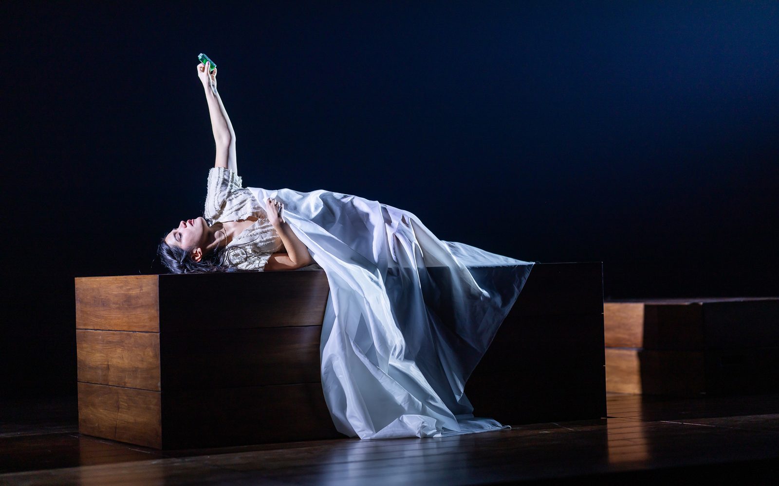 Emilia Suárez lies on a wooden platform under a loose sheet and raises her arm, staring at a small vial in her hand.
