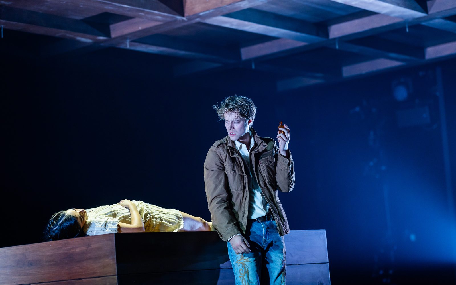 Rudy Pankow in a brown jacket and holding a small vial in his hand, looks down distraught at Emilia Suárez, lying motionless on a wooden platform.