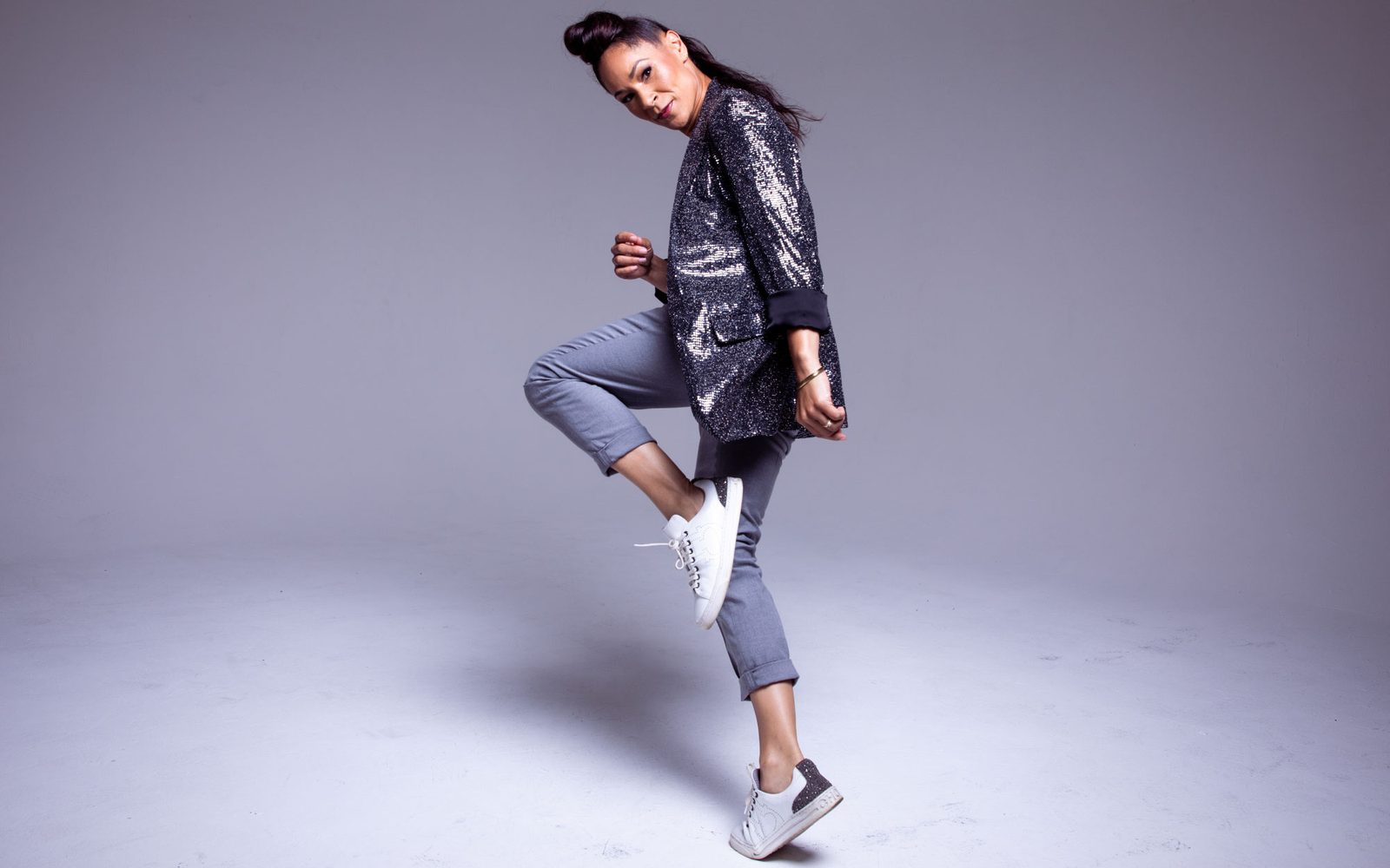 Ayodele Casel in a sparkly jacket lefts one leg in front of a studio background.