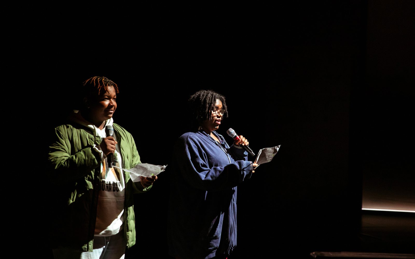 Two students in jackets stand on a darkened stage and read from a page into hand-held mics.