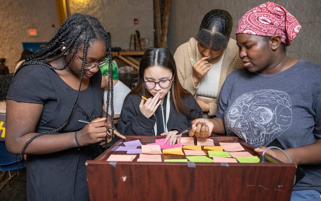 Four students in a dimly lit room gather around a podium with sticky notes spread across it.