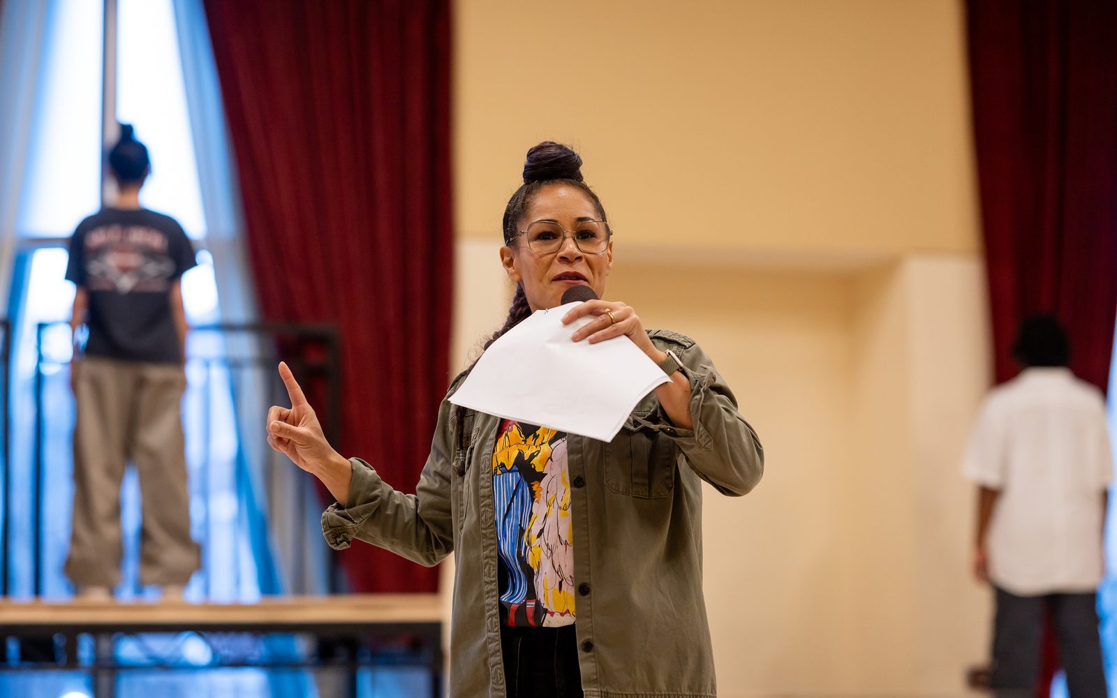 Ayodele Casel in glasses, a high bun, and a brown jacket, speaks into a hand-held microphone while also holding a piece of paper.