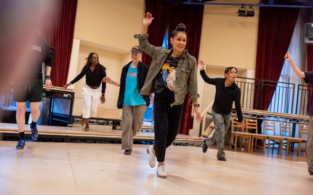 Ayodele Casel in a brown jacket and black jacket raises one arm as she taps in the center of the rehearsal stage, with the other performers around her dancing in unison.