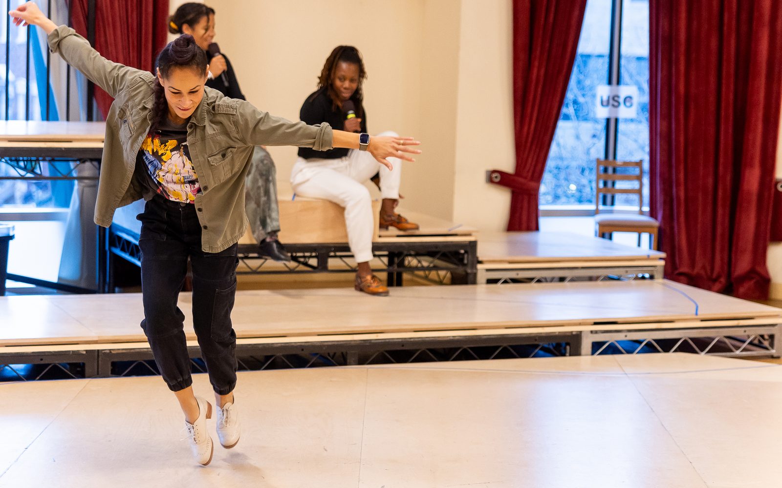 Performer Ayodele Casel in a brown jacket and black pants lifts up on her toes and stretches her arms out. In the background, Ki’Leigh Williams and Quynn L. Johnson sit on a riser and speak into hand-held mics.