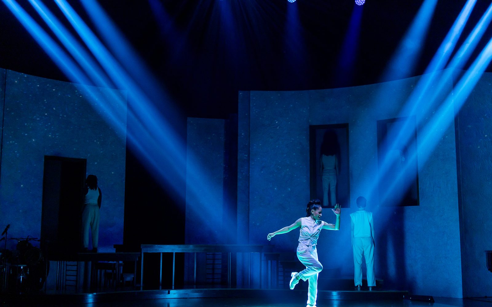 Ayodele Casel in a white vest, pants, and tap shoes, dances center stage lit by multiple spotlights, while other members of the cast face the wall of the set behind her.