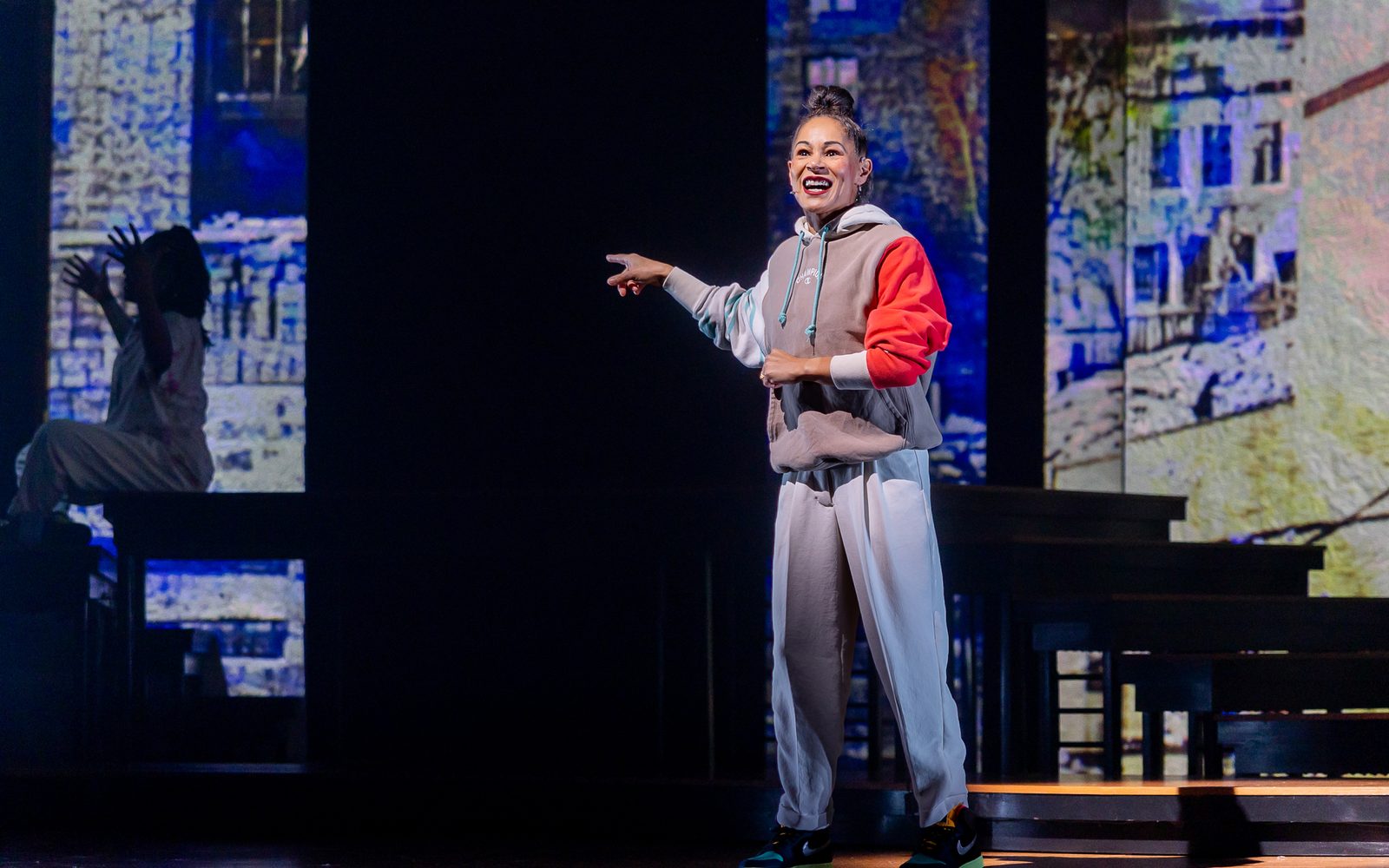 Ayodele Casel in a grey and red sweatshrit, loose white pants, and sneakers, laughs and points to her right.