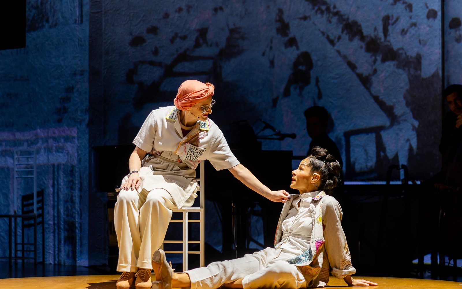 Liberty Styles in a white outfit and a pink turban, sits in a chair and reaches down to touch the shoulder of Ayodele Casel, who sits on the ground in a white outfit with one leg outstretched looking up at them.