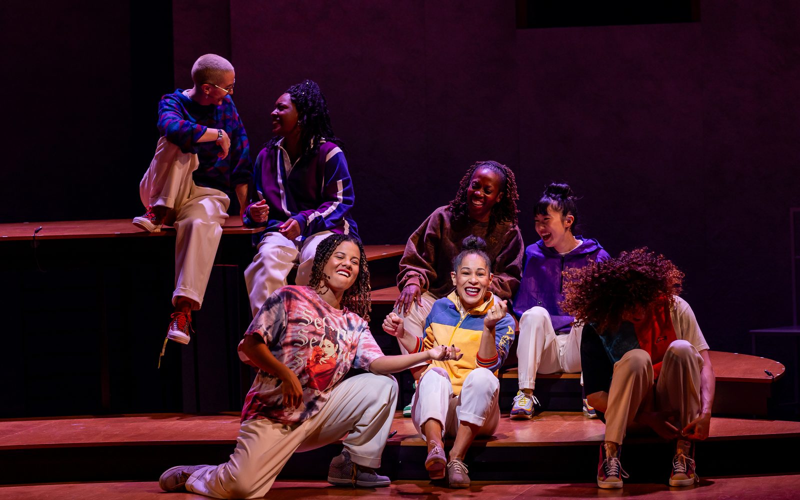 The cast in sweatshirts and loose, white pants, sit gathered on steps, with Ayodele in a blue and yellow track jacket at the center. Ki’Leigh Williams in a pink tie-dye, Selena graphic T-shirt plays an air guitar.