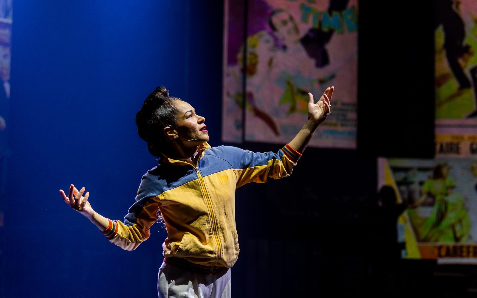 Ayodele Casel in a blue and yellow track jacket strikes a pose, her arms raised on either side of her and her face and updo hairstyle in profile.