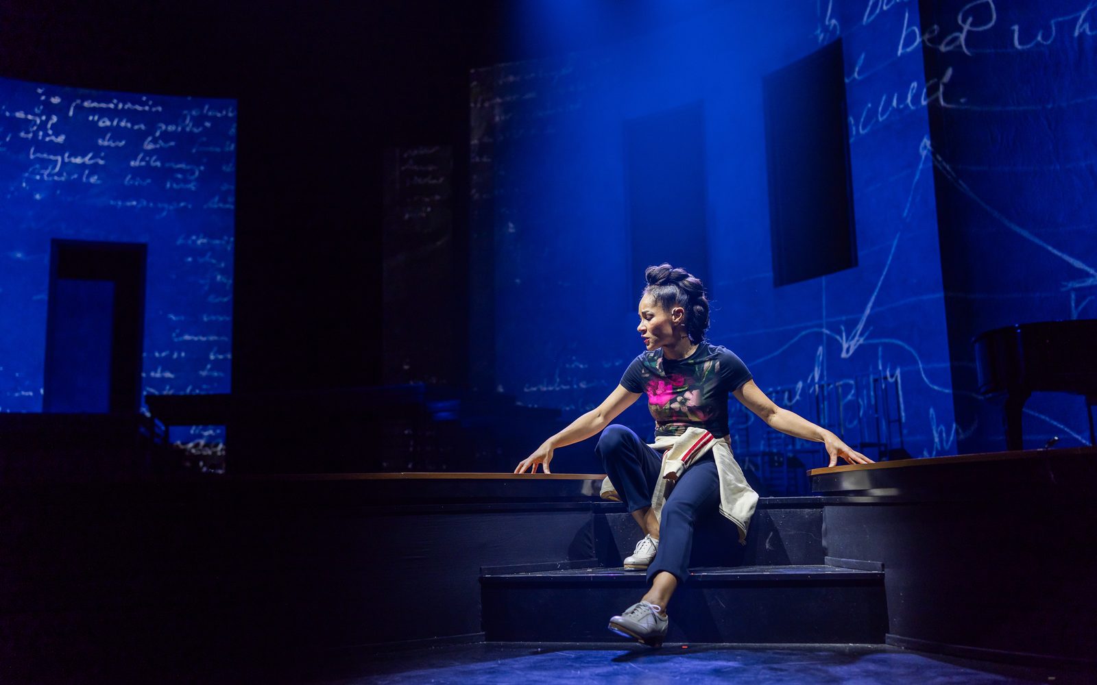 Ayodele Casel in a black graphic T-shirt and navy capri pants sits on steps, extending her arms to rest her fingertips on the floor on either side of her.