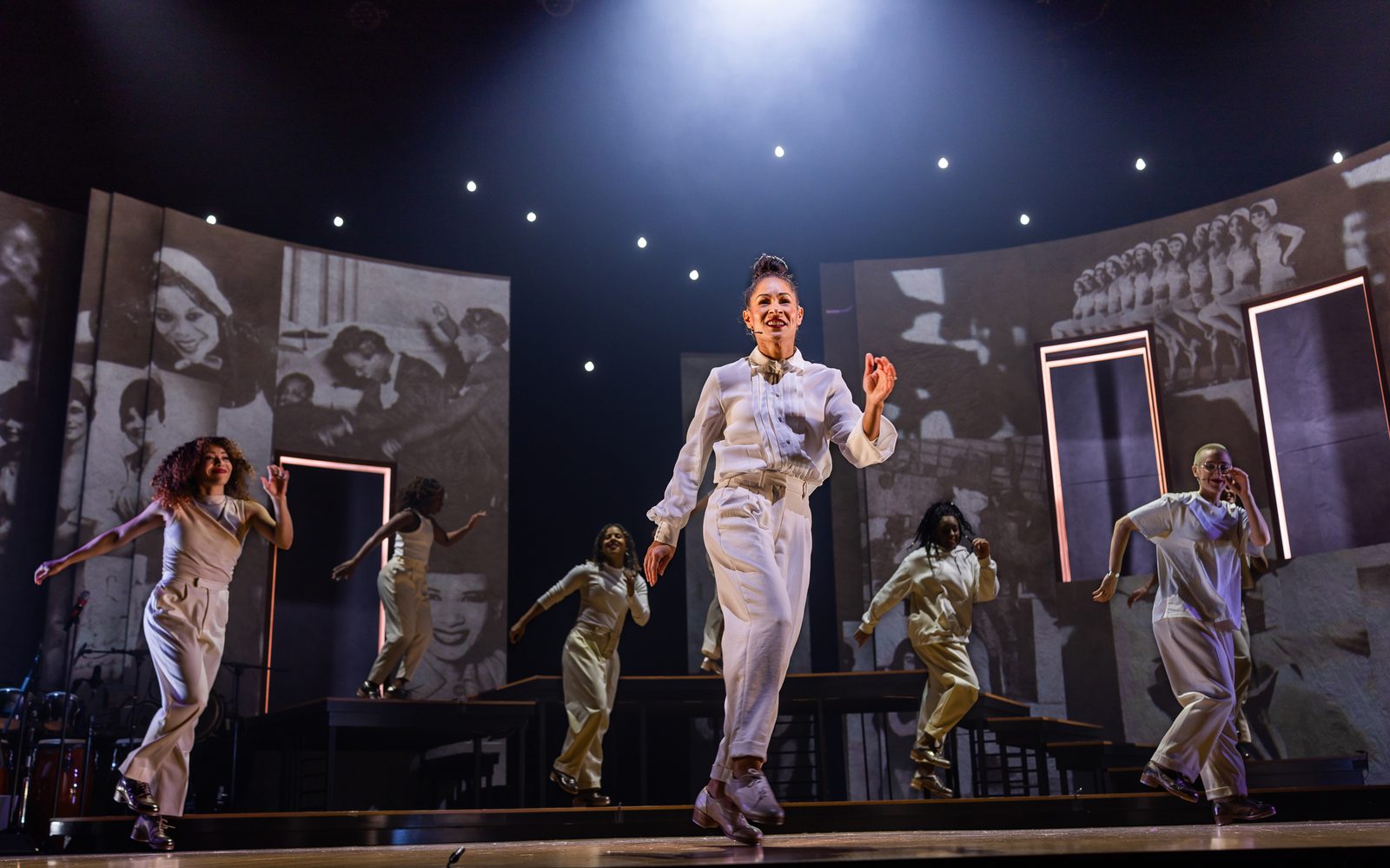 Ayodele Casel in a white tux shirt and slacks dances center stage underneath a spotlight and encircled by the rest of the cast, also dancing in white outfits, while black-and-white historical images of Black tap dancers are projected onto the walls of the set.
