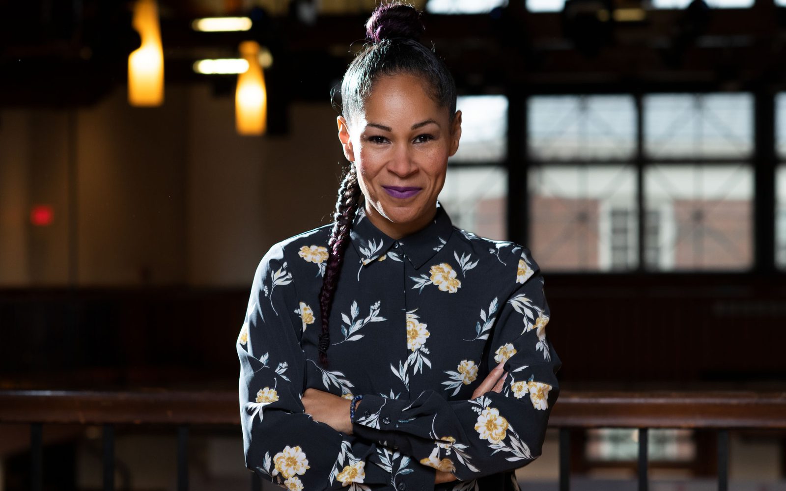 Ayodele Casel in a dark floral blouse with her braid falling over her shoulder crosses her arms and smiles straight into the camera.