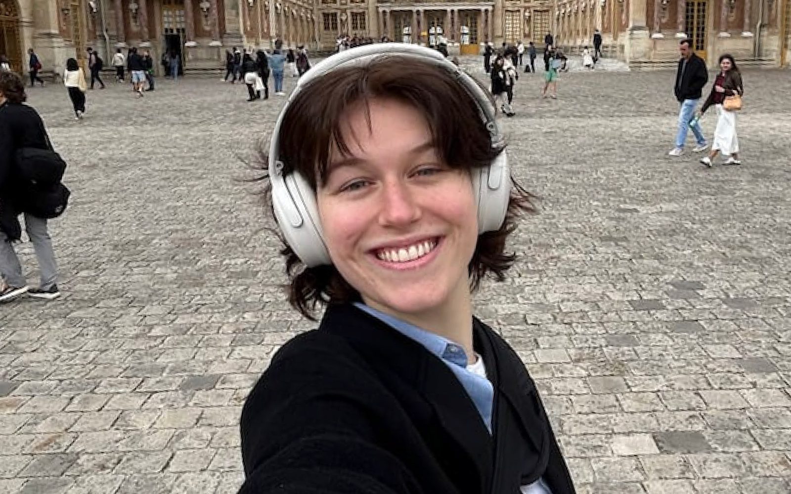 Annaliese Wilbur in a black jacket and large white headphones smiles broadly on a cobblestone street.