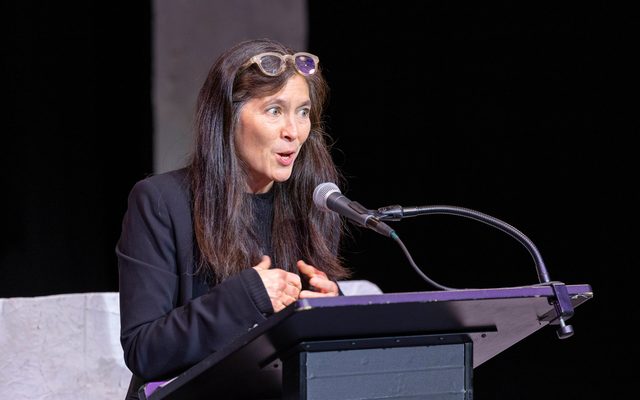 Diane Paulus in a dark navy suit jacket and with glasses atop her head, speaks into a microphone at a podium.