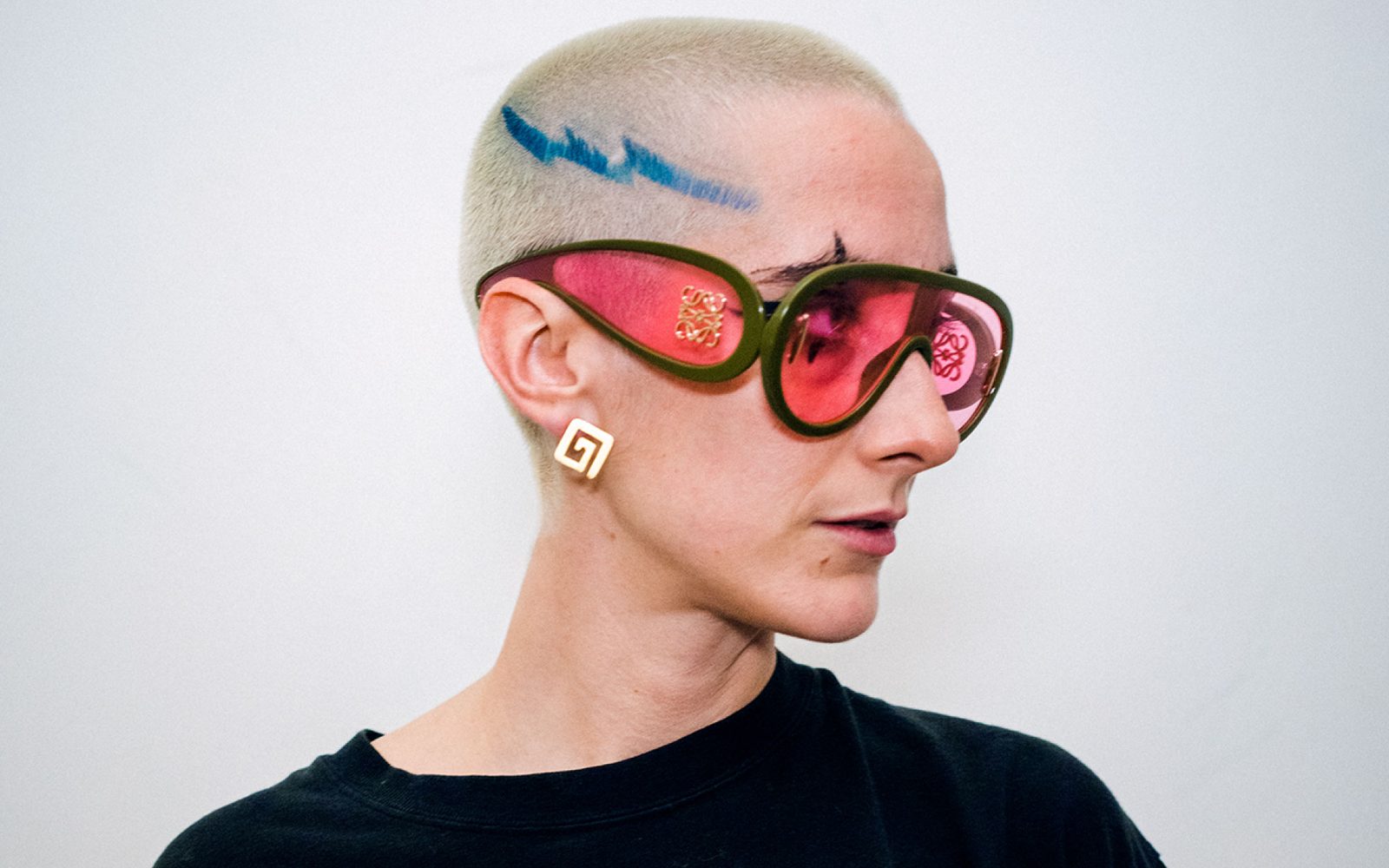 Liberty Styles in profile with buzzed blonde hair with a blue lightning bolt shaved in, oversized red glasses, and a square gold earring.