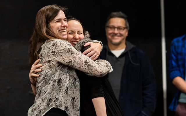 Shana Cooper in a beige blouse with a dot pattern hugs Kate Hamill, while others look on in the background.
