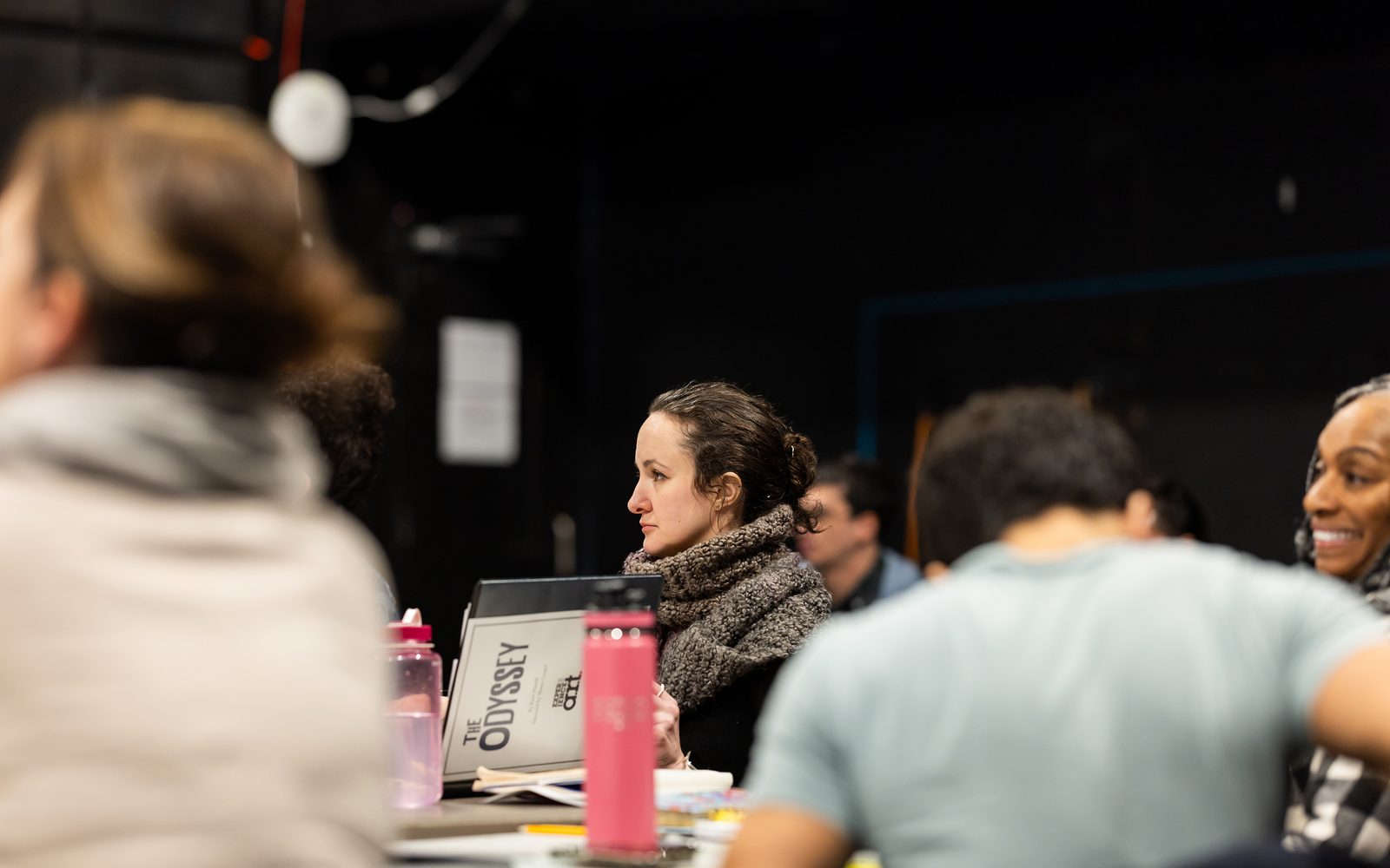 Seen in the gap between other people, Kate Hamill sits at a table in a large grey knit scarf and opens her script.