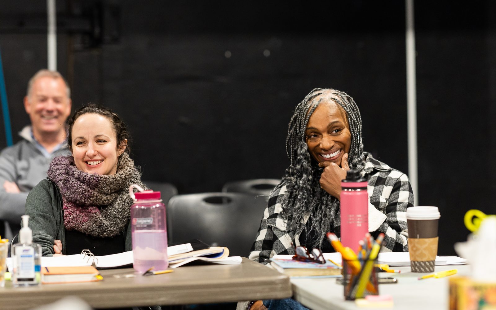 Nike Imoru sitting at a table in a black-and-white plaid shirt and silvery-gray box braids smiles broadly, with Kate Hamill in a large knit scarf sits on the left.